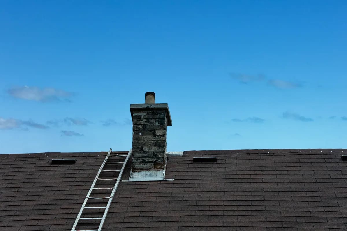 A ladder resting on a shingle roof below a stone chimney
