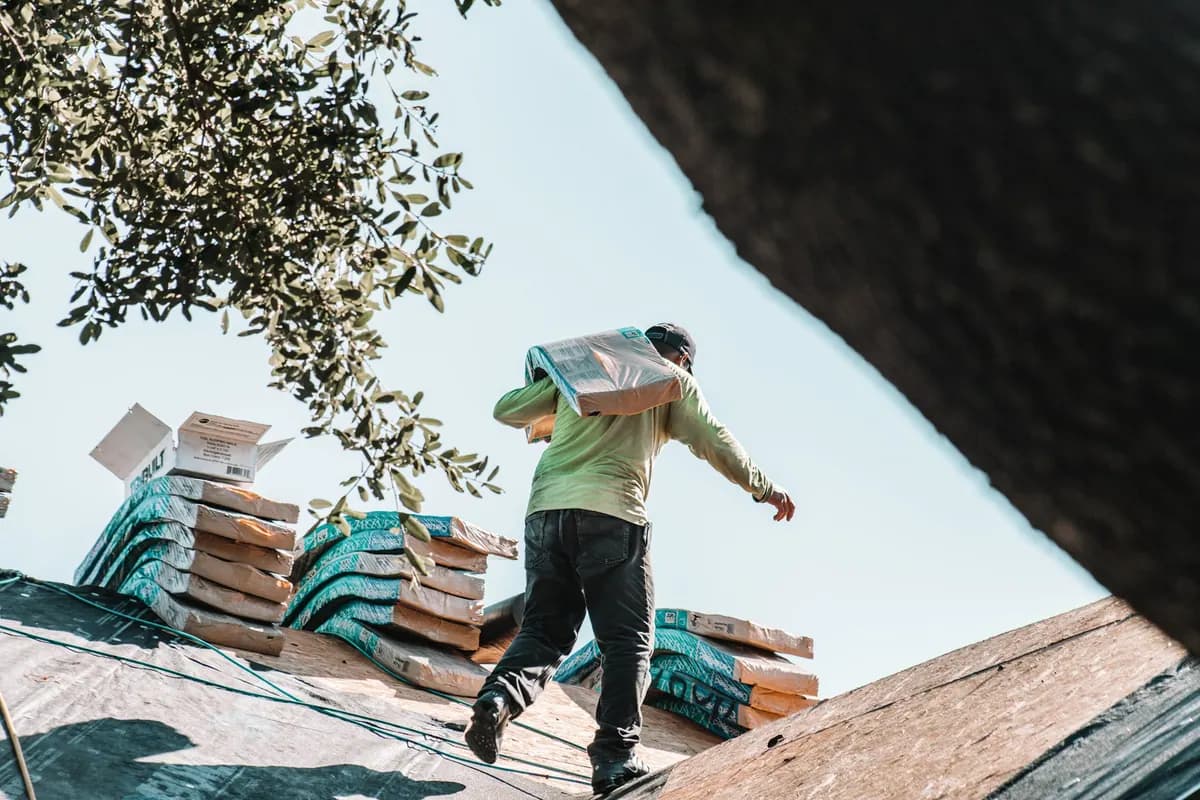 A roofer carrying a bundle of new shingles across a residential roof