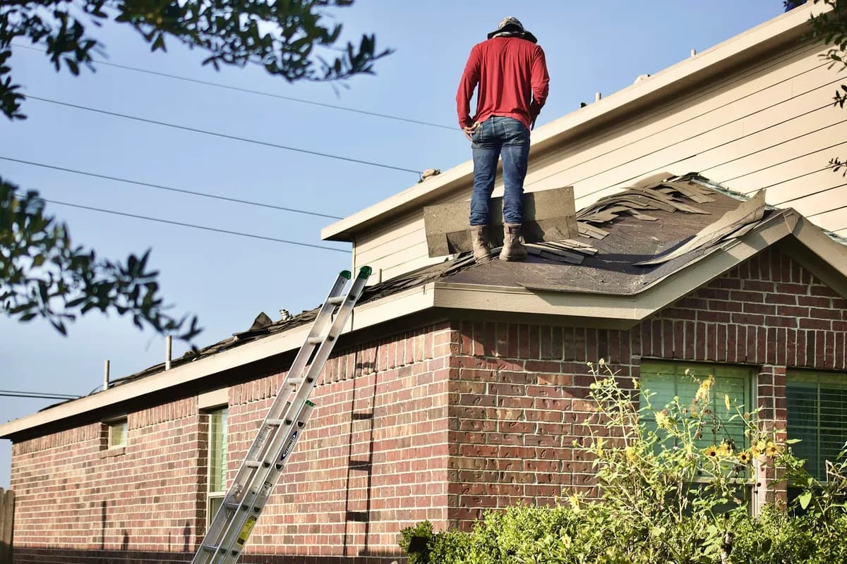 A worker standing on a roof partway through a shingle tear-off, with a ladder against the eave
